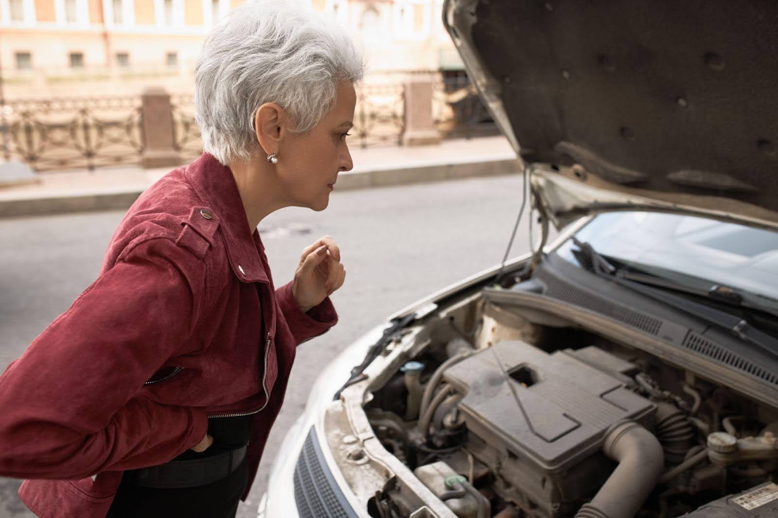 Na imagem, uma mulher idosa de jaqueta vermelha em frente a um carro branco com capô aberto observa o zinabre da bateria.
