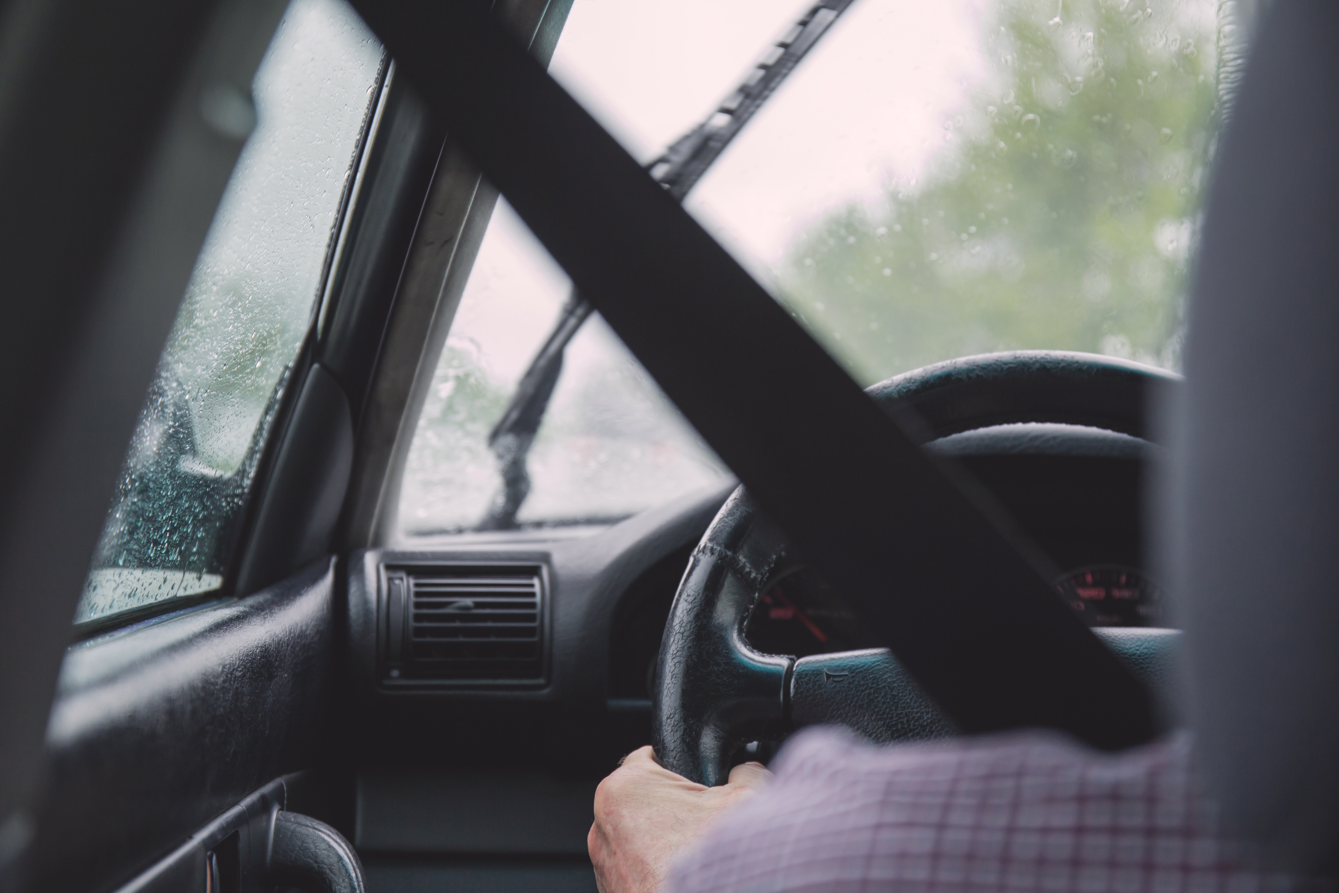 Imagem vista de trás do banco do motorista de um homem dirigindo um carro em dia chuvoso. Ele também usa cinto de segurança.