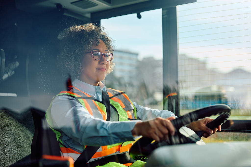 Podemos ver, na imagem, uma motorista dirigindo um ônibus, uniformizada e refletida pelo vidro dianteiro do veículo.