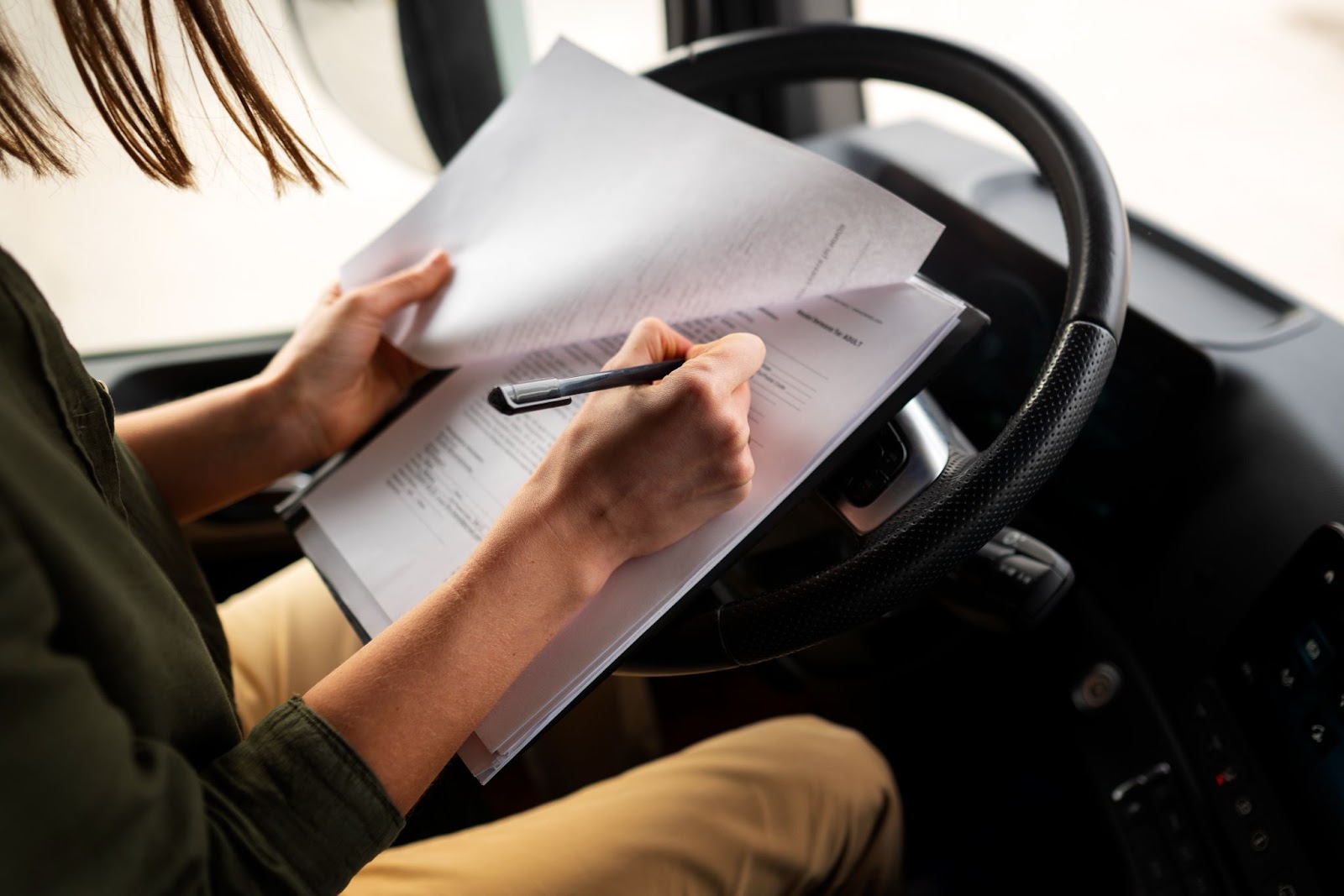 Mulher sentada no banco de motorista apoia uma prancheta no volante enquanto preenche um formulário.