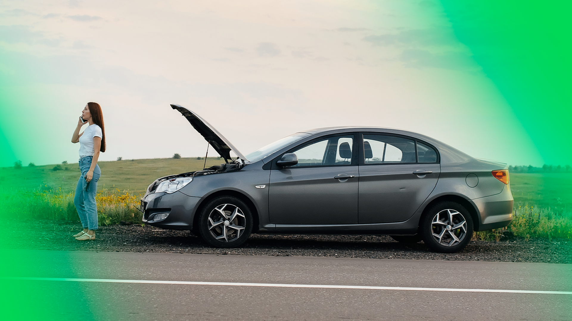 Imagem mostra carro no acostamento de estrada, comcapô aberto, e mulher em pé do lado de fora, à frente do carro, usando celular para chamar ajuda mecânica ou guincho.