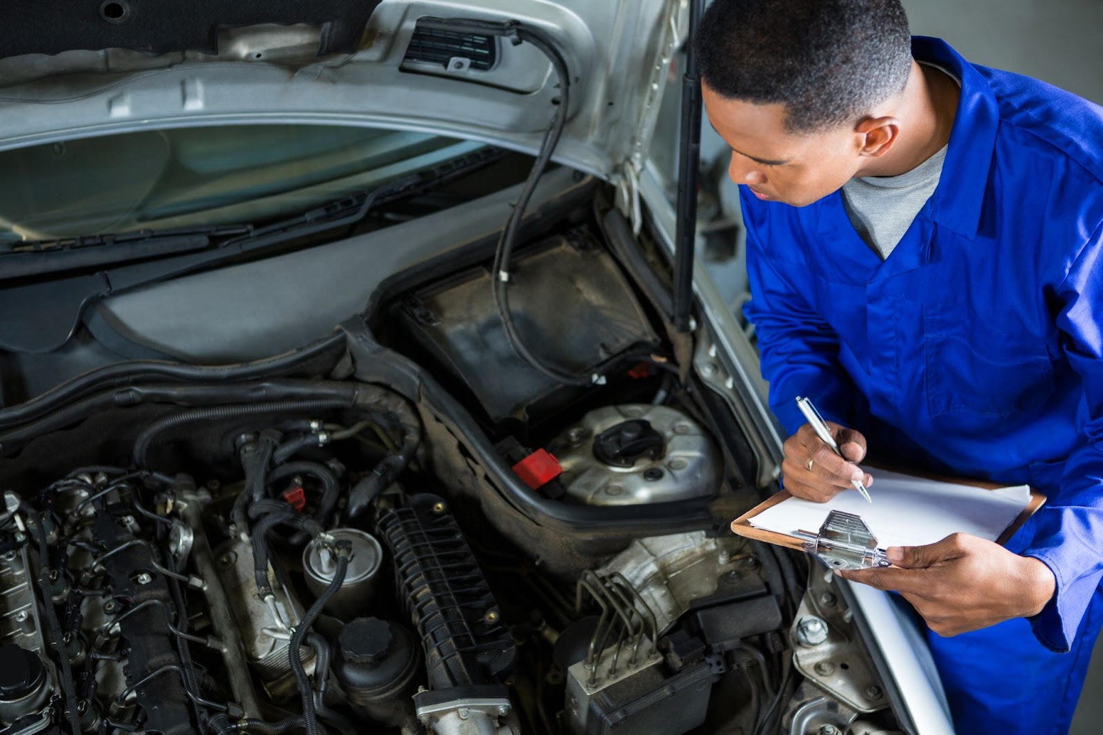 Um mecânico de jaleco azul faz a manutenção preventiva observando o motor de um carro enquanto anota em uma prancheta.