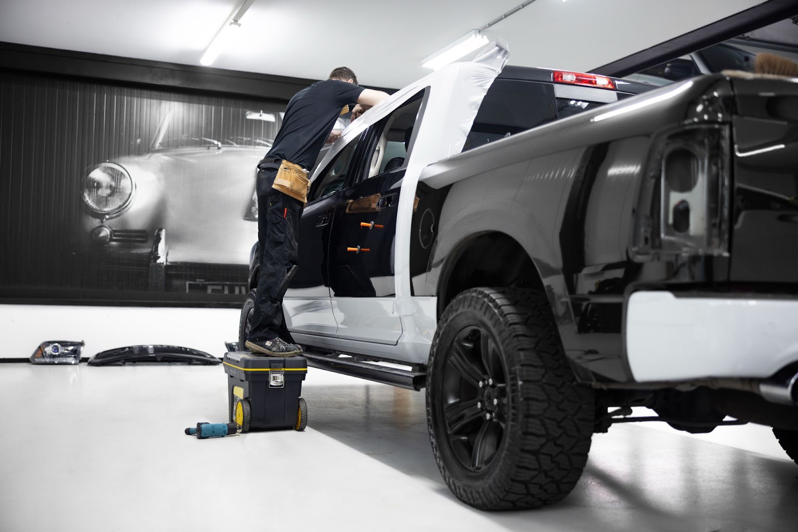 Na imagem, há um homem de macacão em pé sobre uma caixa de ferramentas fazendo modificações em uma caminhonete preta que vai precisar de um certificado de segurança veicular.
