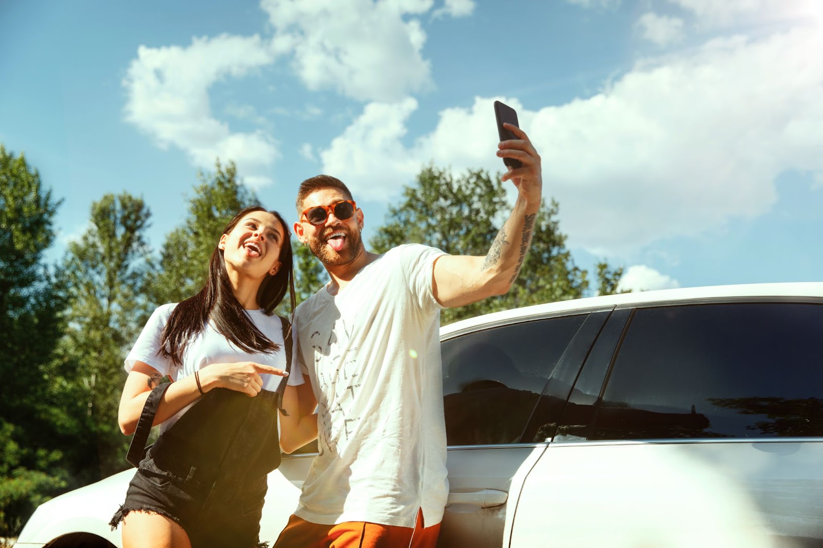 Um casal está de pé junto a um carro branco, capturando uma selfie para registrar a viagem de carro pelo Brasil, desfrutando da luz do dia.