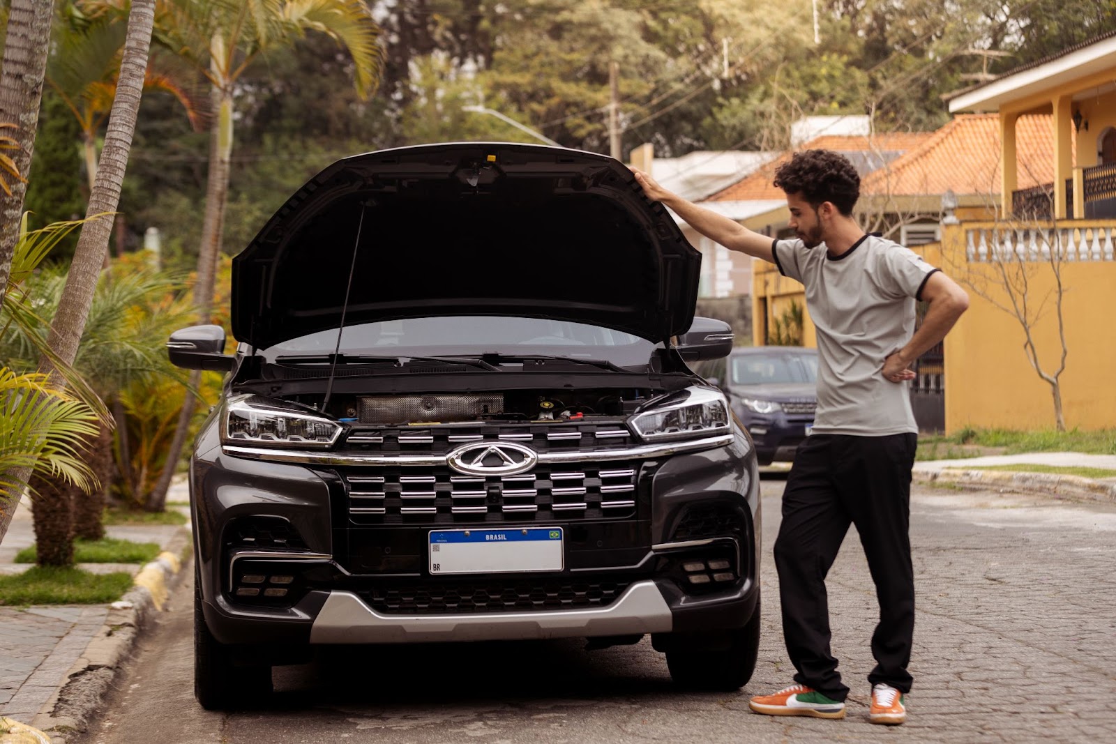 Um homem de camisa branca e calça preta está ao lado de um carro estacionado com o capô aberto, ele está com uma mão apoiada na cintura e observa o carro com expressão de dúvida.