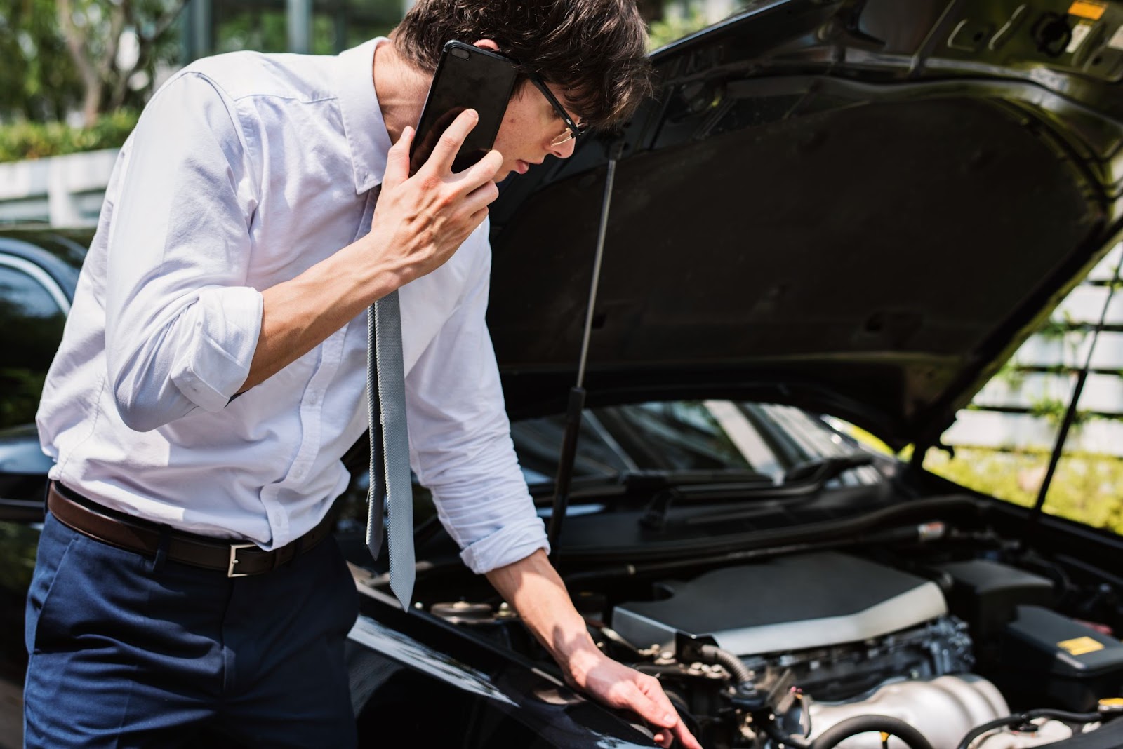 Um homem de camisa social branca e gravata cinza está ao lado de um carro estacionado com o capô aberto, ele observa a bateria enquanto fala ao telefone.