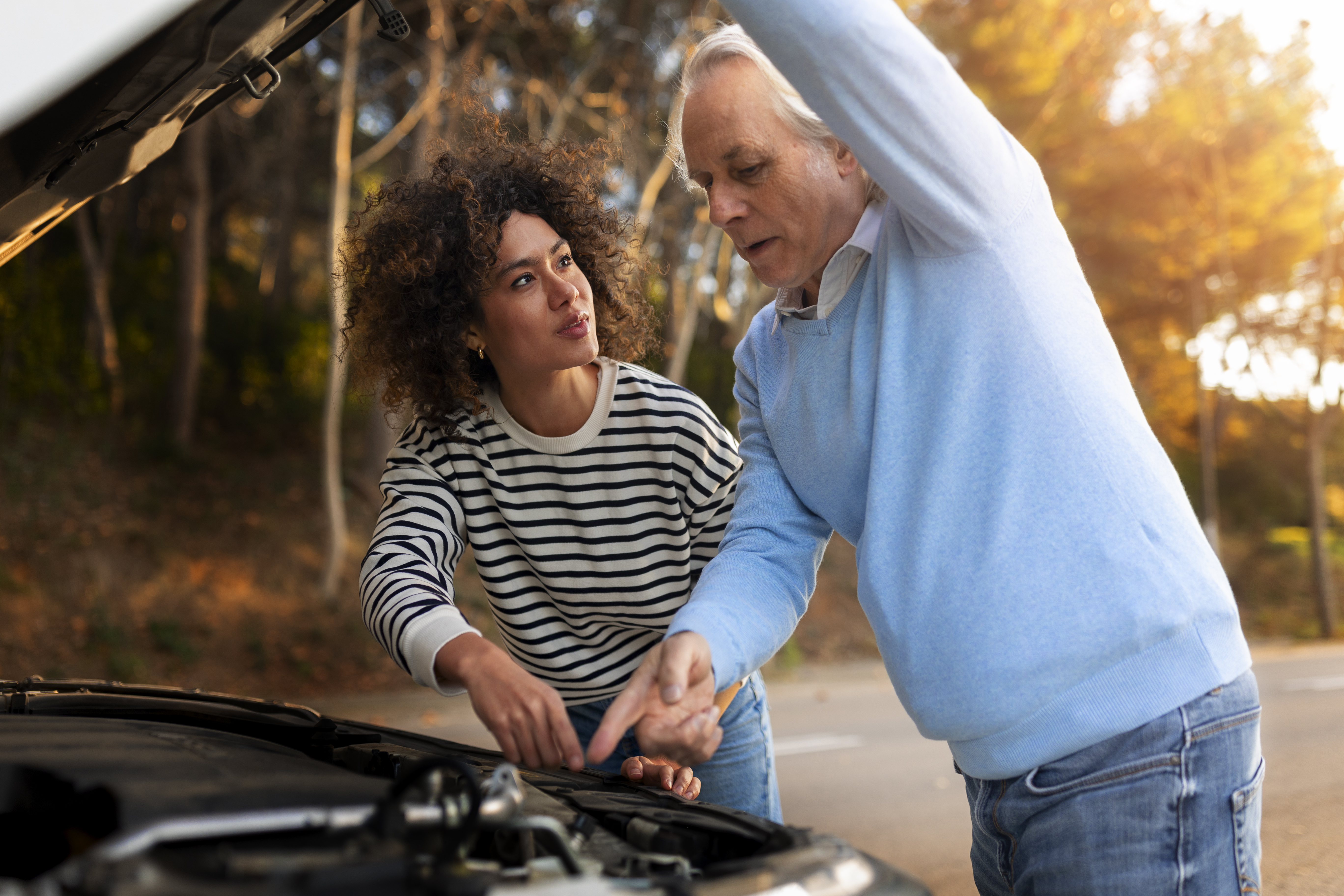 Um homem de suéter azul claro abre o capô de um carro enquanto conversa sobre os cuidados com o conversor de torque com uma mulher de cabelos cacheados e suéter listrado.