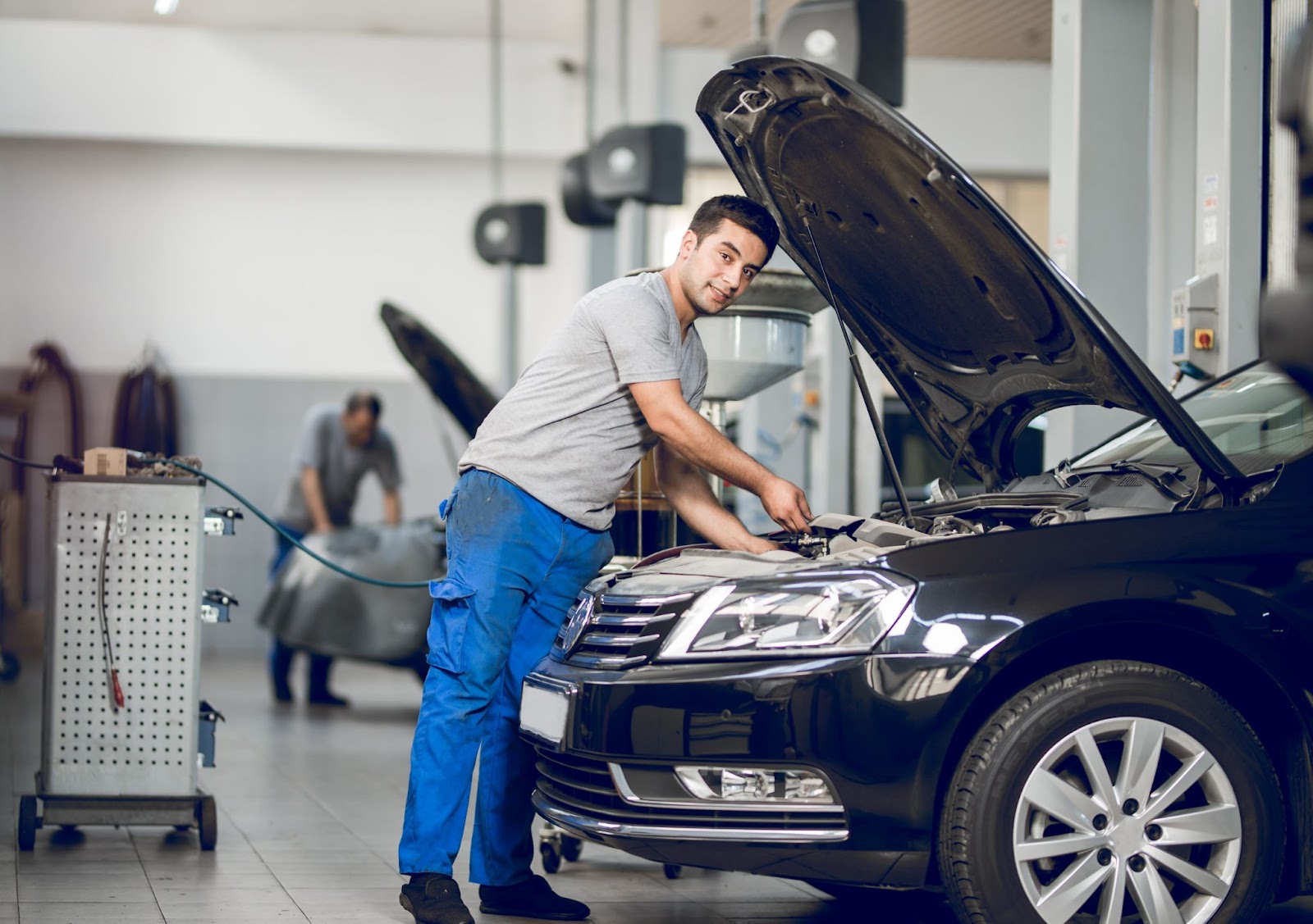 Um mecânico está usando uma camiseta cinza e calça azul enquanto verifica o sistema de injeção eletrônica de um carro com o capô aberto em uma oficina.