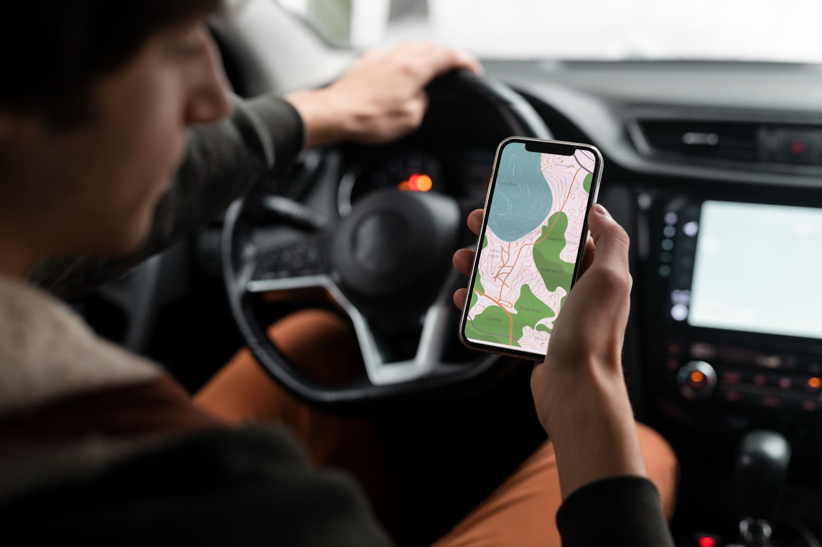 Um motorista está dentro do carro pronto para dirigir na chuva com uma mão no volante e segurando um celular, que exibe o GPS para auxiliar na navegação.