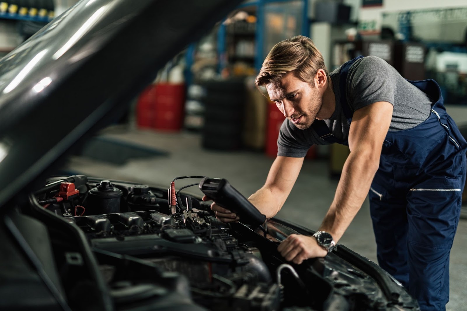 Um técnico automotivo vestindo um macacão azul está usando um equipamento de teste para avaliar a carga da bateria do carro. Ele está inclinado sobre o veículo com o capô aberto, segurando o equipamento na mão direita.