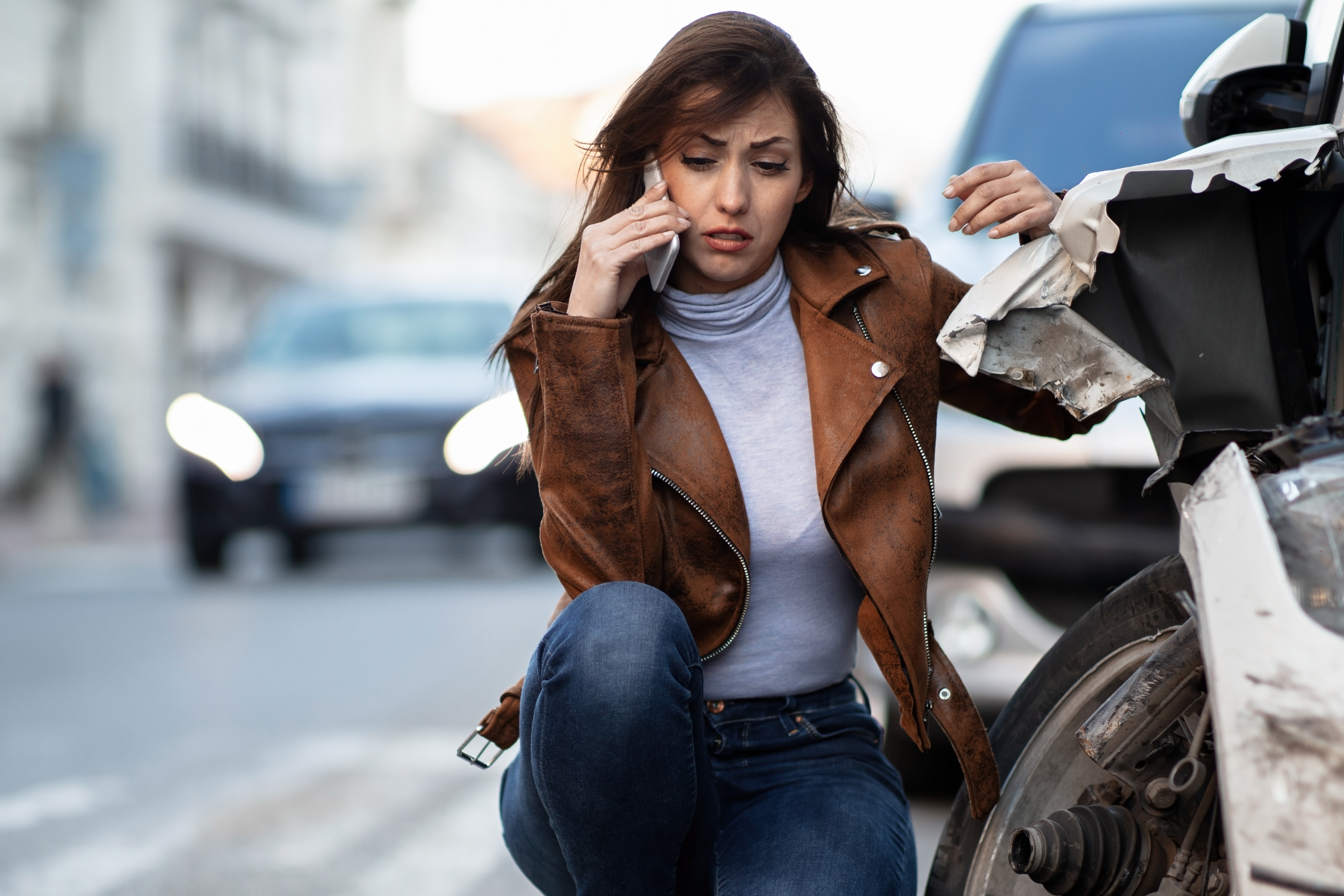 Uma mulher de jaqueta de couro marrom está abaixada ao lado de um carro branco que sofreu colisão e está com a junta homocinética quebrada. Ela fala ao telefone com expressão triste.