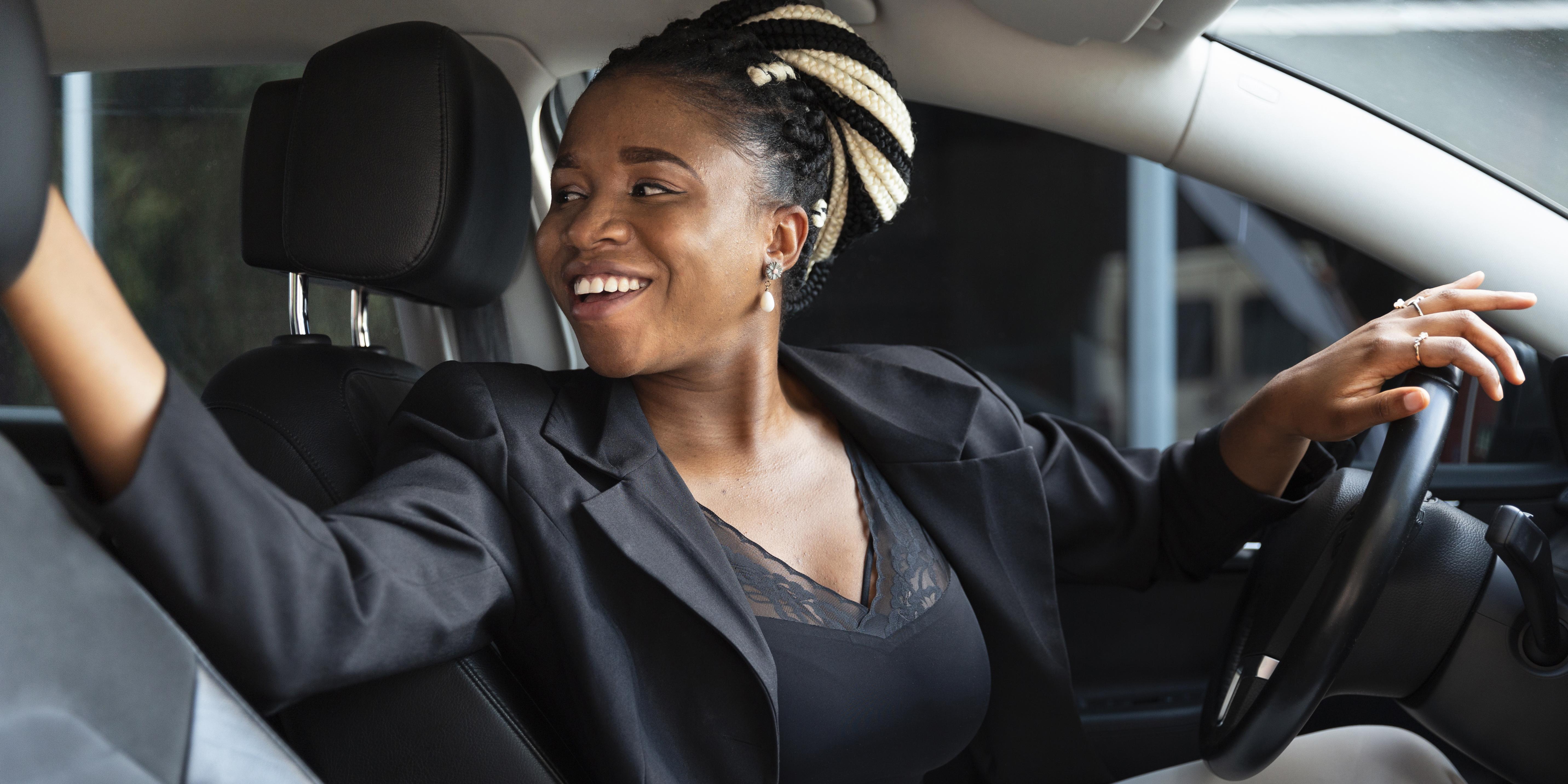 Uma mulher de roupa preta e cabelo trançado está sentada ao volante de um veículo e olha para trás sorrindo enquanto manobra o veículo habilidosamente.