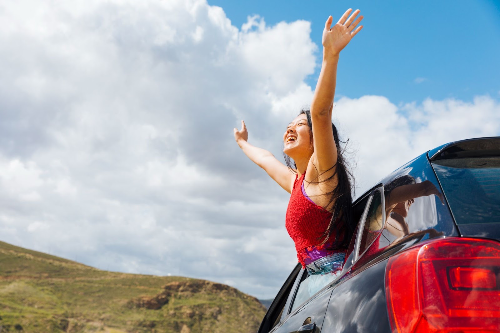 Uma mulher em uma viagem de carro pelo Brasil, encontra-se com metade do corpo para fora da janela do veículo, estendendo os braços e exibindo um sorriso enquanto aprecia a paisagem natural durante o dia