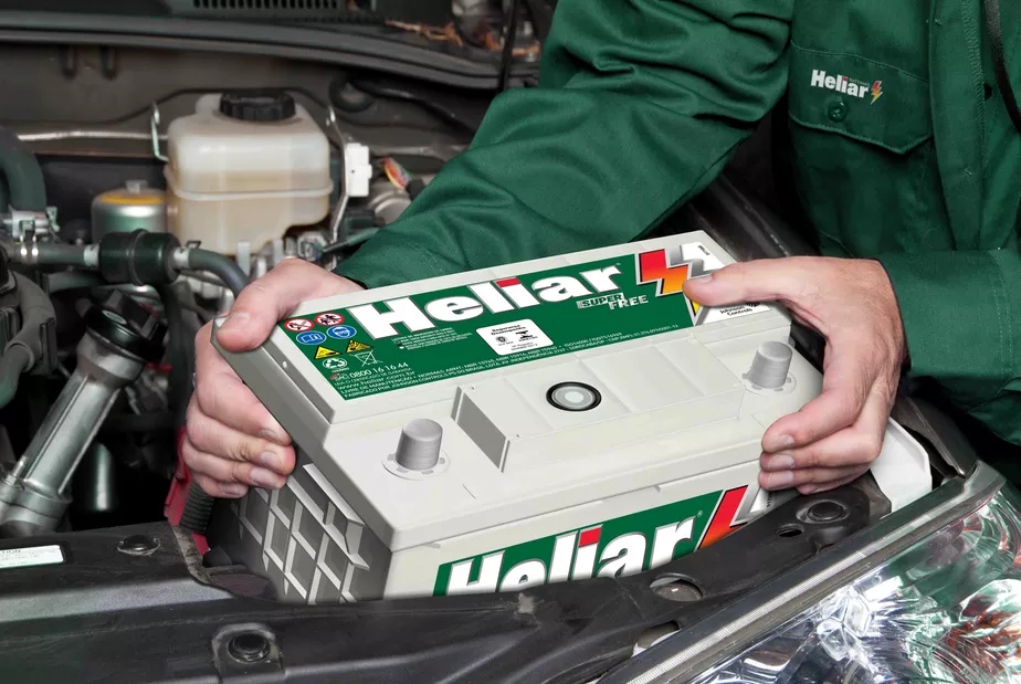 Close-up de mecânico posicionando bateria Heliar para o funcionamento de um carro. Ele veste uniforme verde com a marca Heliar estampada no bolso da camisa.