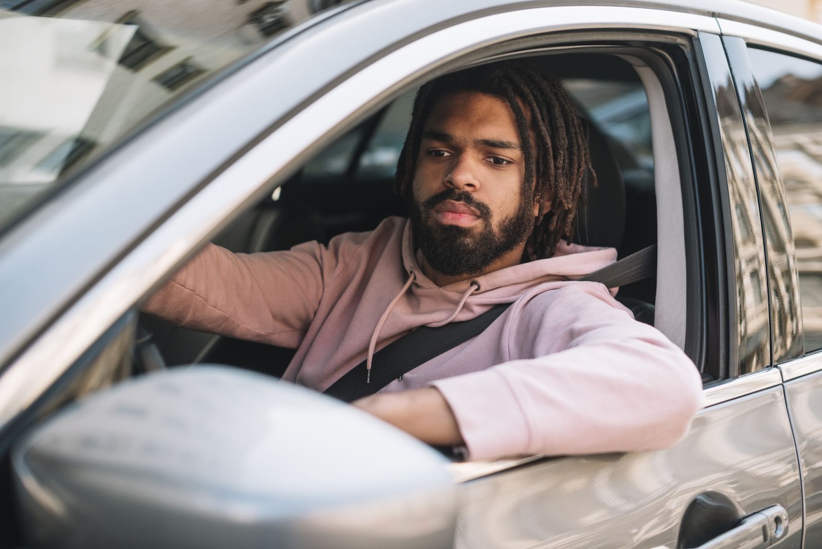 Homem dirigindo preocupado com o alinhamento e balanceamento de seu carro. Ele veste moletom rosa claro e apoia o braço na janela.