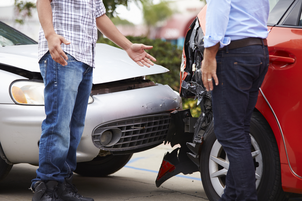 Imagem de um acidente de carro, em que um veículo prata bateu na traseira de um veículo vermelho. Dois homens estão parados em frente aos carros.