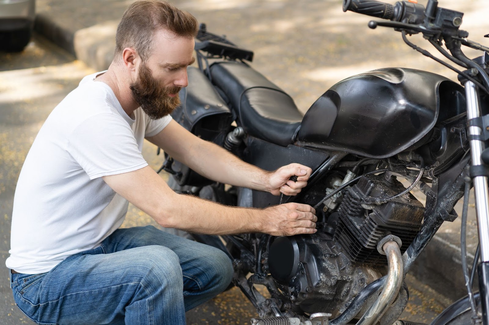 Homem de camiseta branca e calça jeans agachado em frente a uma moto e checando a bateria. 