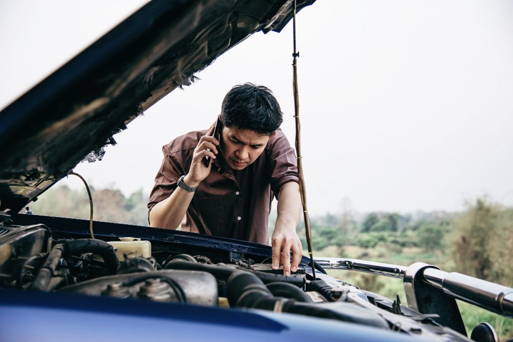 Homem parado na estrada com o capô do carro aberto enquanto fala no celular para resolver o problema do superaquecimento do motor.