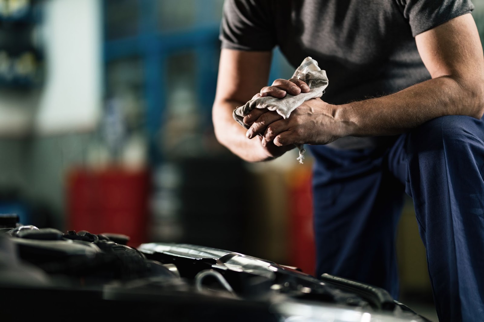 Close-up de mecânico em frente a um carro com capô aberto se preparando para consertar superaquecimento do motor enquanto limpa as mãos com um pano.
