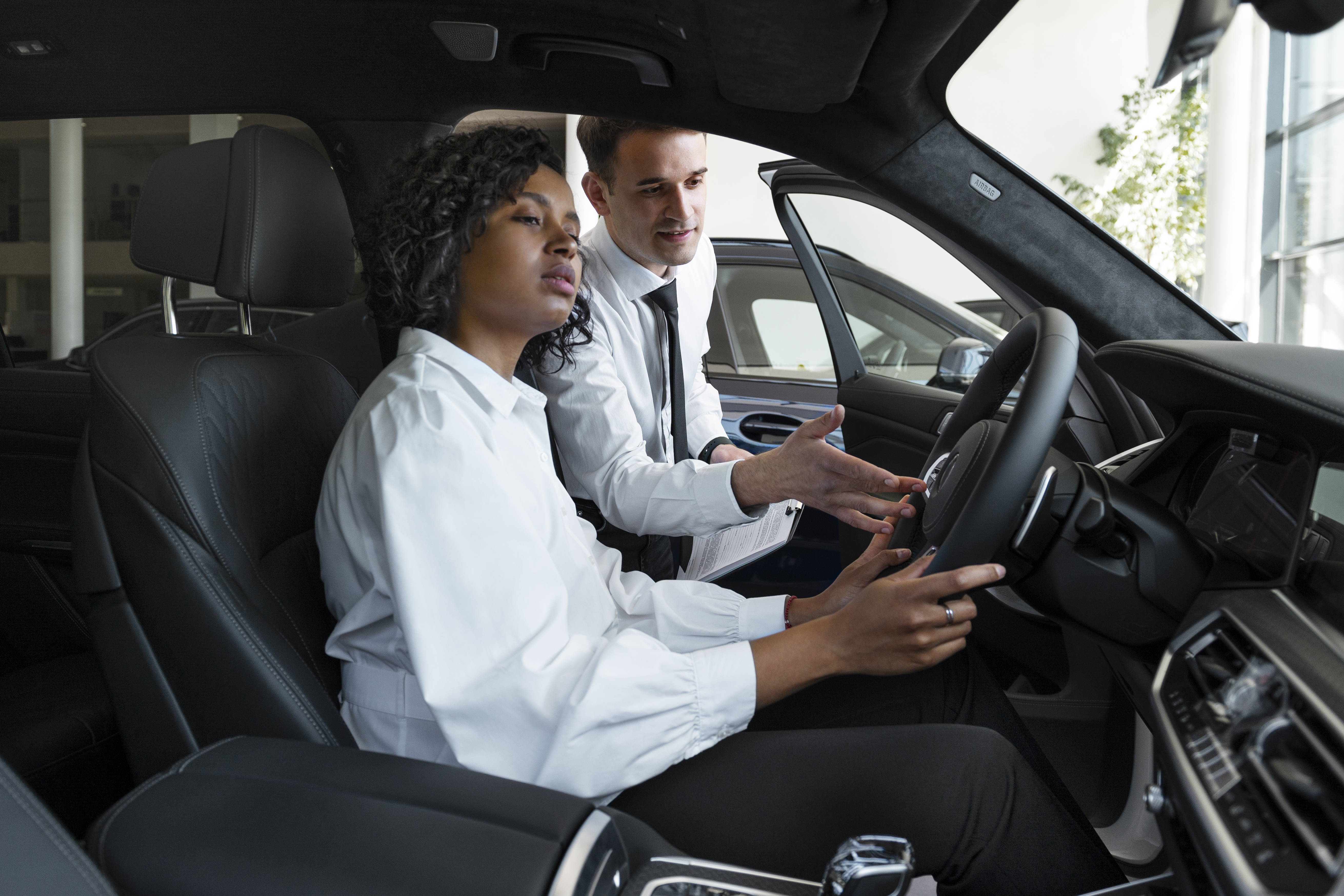 Mulher jovem sentada no banco do motorista com as mãos no volante. Ao seu lado, um vendedor segura uma planilha e explica qual é a melhor direção: elétrica ou hidráulica.